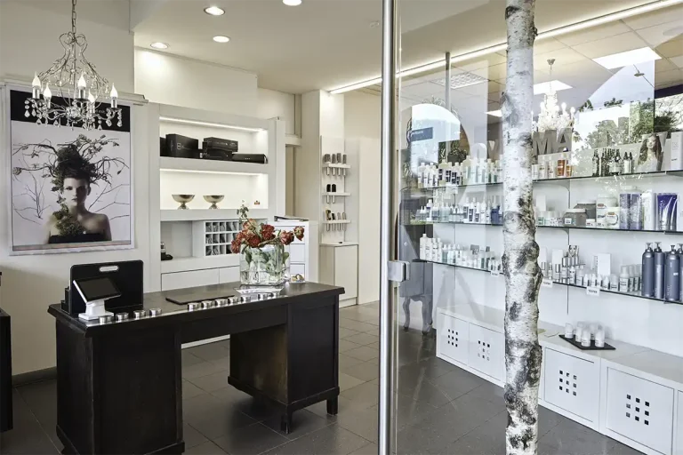 Das Bild zeigt den Friseursalon mit einem dunkelbraunen, alten Holztisch auf der linken, sowie ein Regal mit Pflegeprodukten auf der rechten Seite