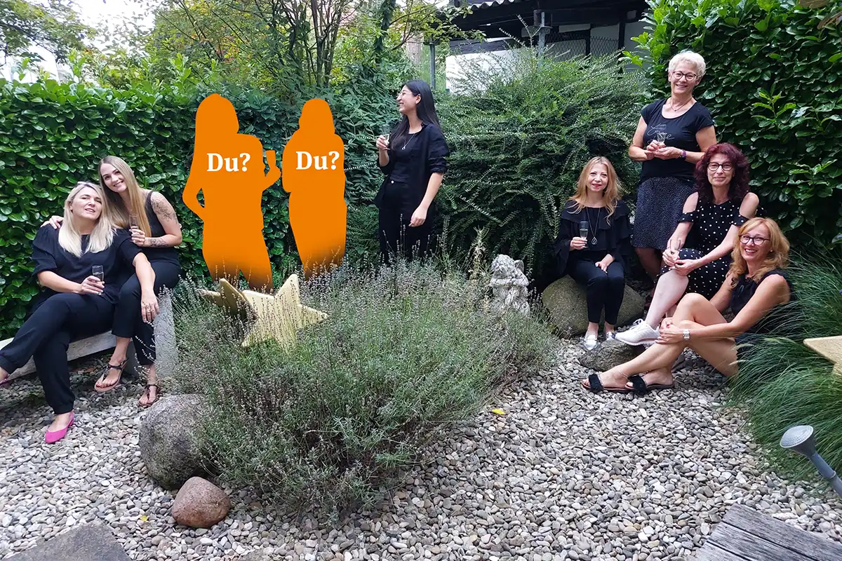 Gruppenbild mit Mitarbeiterinnen von Overmannn Frisuren, die mit einem Sektglas in der Hand neben zwei Mitarbeiter-Platzhalter mit der Aufschrift „Du?“ im saloneigenen Garten stehen und in die Kamera schauen
