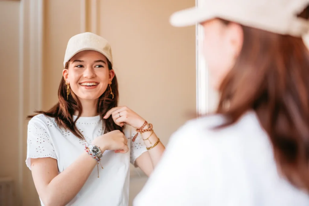 Model, das im weißen Outfit und weißer Schildkappe lächelnd vor dem Spiegel steht und mit ihren Händen die Haare ihres schulterlangen braunen Toupim-Eigenhaarband arrangiert