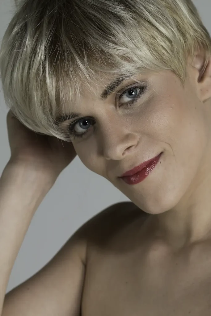 Model, das sich mit der Belle-Madame-Perücke „Coco Mono SF“ in Champagner Root ins Haar fasst und mit geneigtem Kopf die Kamera blickt
