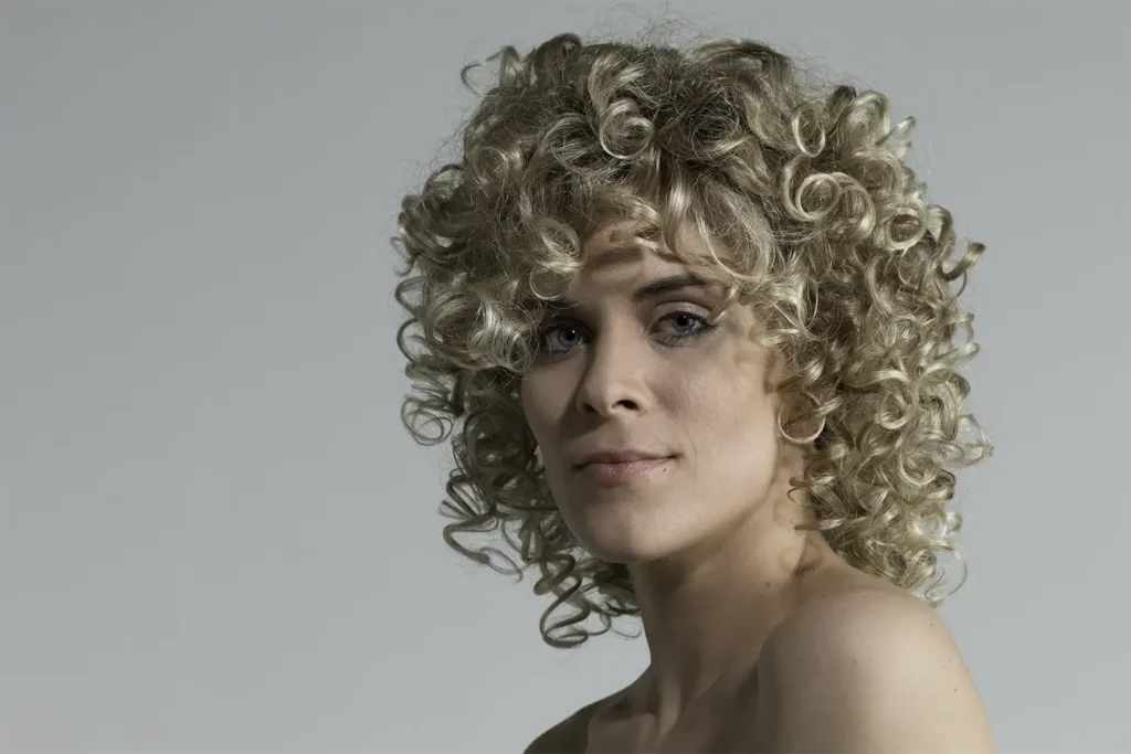 Model, das mit einer blonden, lockigen Perücke von Ellen Wille und seitlich gedrehtem Körper in die Kamera blickt