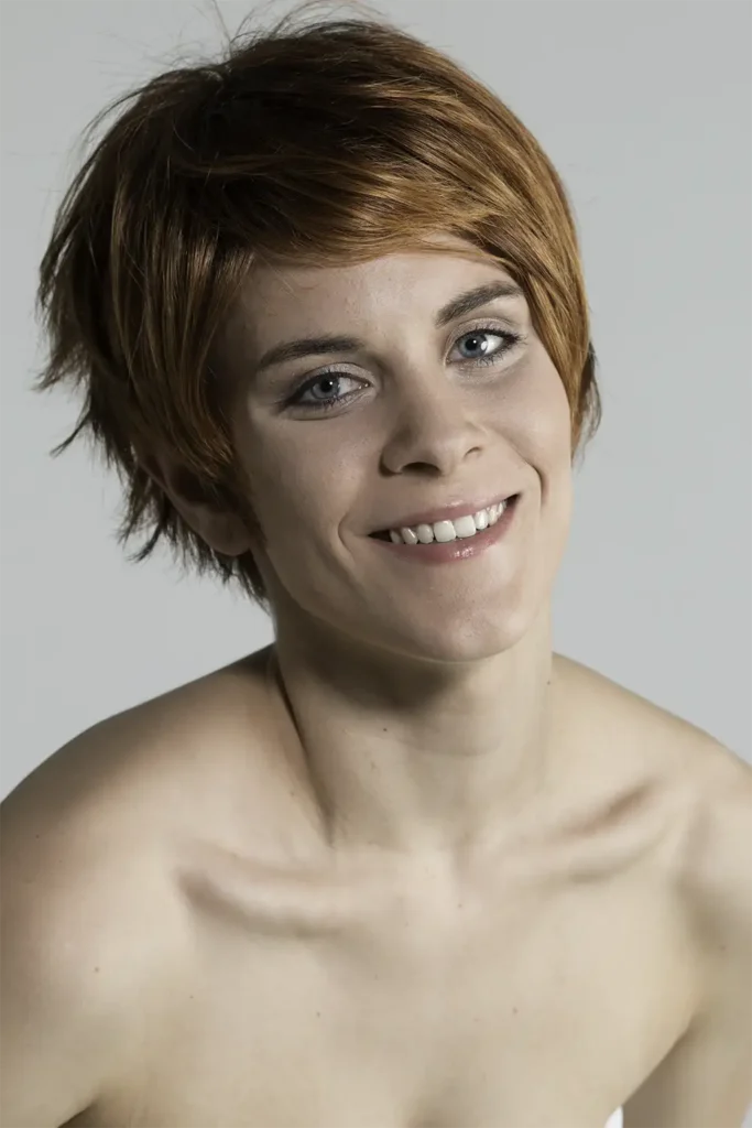 Model, das mit Belle-Madame-Perücke „Tilda Soft“ in Terracotta-Mix Root mit leicht geneigtem Kopf die Kamera blickt